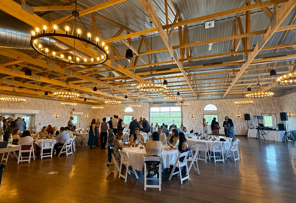 Wide shot of the entire wedding venue. Warm-toned wooden floors, rustic, warm lighting, round tables and white chairs. Big windows can be seen in the back