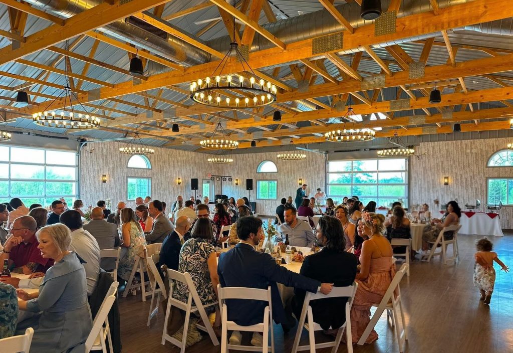 Lots of people sitting at round tables with white chairs in a venue with wooden floors and soft warm lighting.